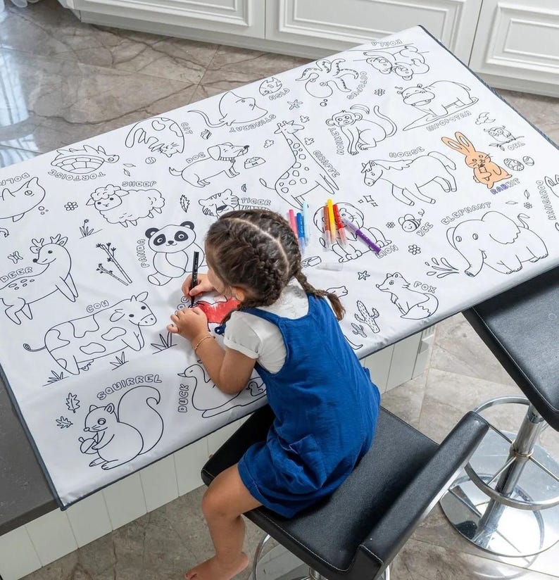May include: A white tablecloth with black outlines of various animals, including a deer, cow, squirrel, duck, panda, fox, elephant, giraffe, and more. A child is coloring the tablecloth with markers.