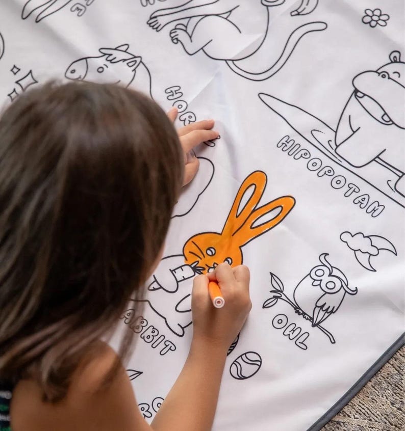 May include: A child colors a white fabric mat with an orange marker. The mat features black and white illustrations of animals, including a rabbit, an owl, a hippopotamus, and a horse. The words "RABBIT", "OWL", "HIPOPOTAM", and "HORSE" are written next to the animals.