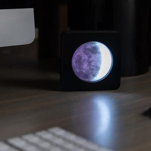 May include: A black square-shaped night light with a moon-shaped design. The moon is illuminated with a white light and has a purple hue on the left side.