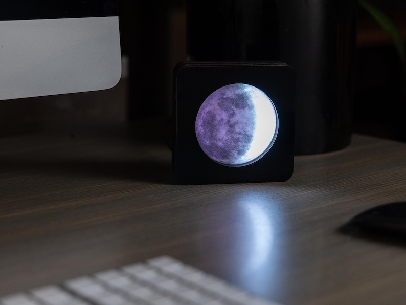 May include: A black square-shaped night light with a moon-shaped design. The moon is illuminated with a white light and has a purple hue on the left side.