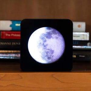 May include: A black square-shaped moon lamp with a white moon image inside. The moon is in a crescent phase and has a purple hue.