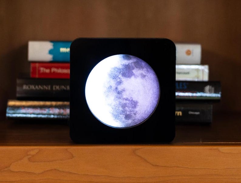 May include: A black square-shaped moon lamp with a white moon image inside. The moon is in a crescent phase and has a purple hue.