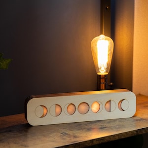 May include: A rectangular light-colored wooden lamp featuring a series of seven circular cutouts that display the phases of the moon. The lamp emits a warm, soft glow from within, highlighting the circular moon phase designs across its front face.