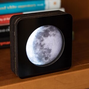 May include: A black wooden box with a clear front panel displaying a detailed image of the moon. The moon is illuminated from behind, creating a soft glow.