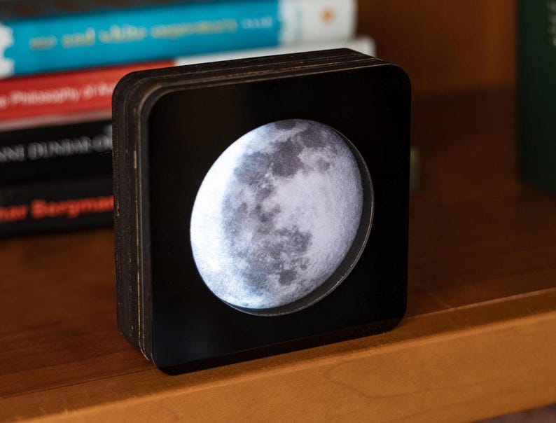 May include: A black wooden box with a clear front panel displaying a detailed image of the moon. The moon is illuminated from behind, creating a soft glow.
