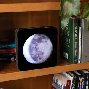 May include: A square, black, illuminated lamp featuring a detailed, circular, high-contrast photographic print of the moon in its waxing gibbous phase, positioned on a wooden shelf between several hardcover books.
