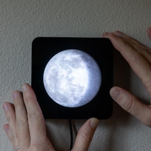May include: A black square-shaped moon lamp with a realistic image of the moon's surface. The lamp is mounted on a wall and is illuminated, casting a soft glow.