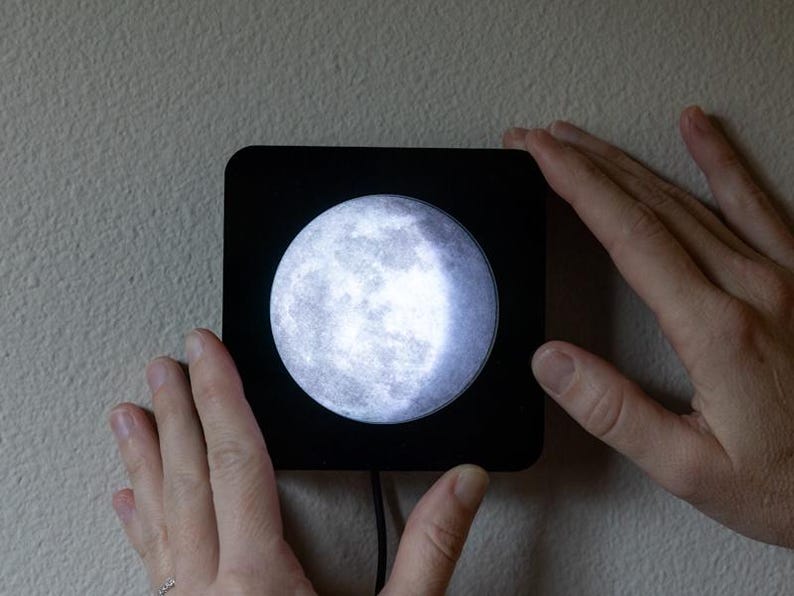 May include: A black square-shaped moon lamp with a realistic image of the moon's surface. The lamp is mounted on a wall and is illuminated, casting a soft glow.