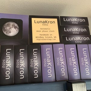 May include: A display of LunaKron MINI automatic moon phase clocks. The clocks are handmade on Whidbey Island, WA. The text "LunaKron MINI" is repeated on each clock.