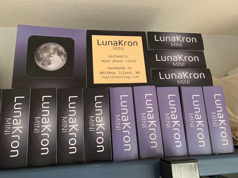 May include: A display of LunaKron MINI automatic moon phase clocks. The clocks are handmade on Whidbey Island, WA. The text "LunaKron MINI" is repeated on each clock.
