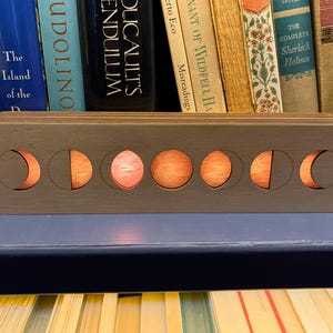 May include: A wooden moon phase lamp in grey. It has six cut-out moon phases that glow orange. The rectangular lamp has rounded edges and sits on a blue surface. The background features a collection of books.