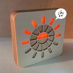 May include: A square, wooden speaker with a sunburst design on the front. The sunburst is made of orange-colored accents. A musical note icon is in the upper right corner. The speaker is made of layered wood.