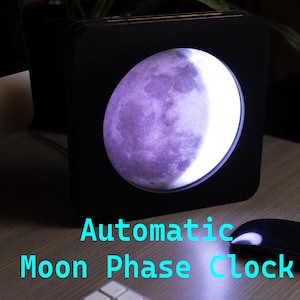May include: A black wooden clock with a circular moon phase display. The moon is illuminated in a purple hue and shows a crescent moon shape. The text "Automatic Moon Phase Clock" is printed below the clock.