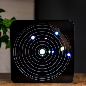 May include: A square black display panel featuring a white concentric circle pattern representing planetary orbits. Several small, glowing, multi-colored LED lights are positioned along these circular paths, mimicking a solar system model.