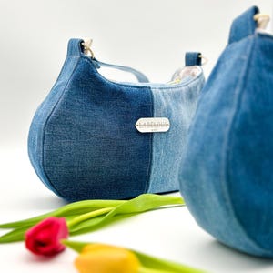 Könnte beinhalten: Zwei Jeans-Handtaschen in Blau mit abgerundeter Form. Jede Tasche hat ein kleines Metallschild mit der Aufschrift "LABELOUS". Eine rote und eine gelbe Tulpe befinden sich im Vordergrund.