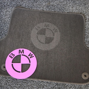 Può includere: Un tappetino per auto grigio con un logo BMW nero e un ritaglio rosa del logo BMW con le lettere "BMW" intorno.