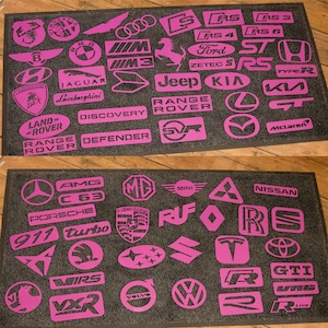Puede incluir: Una alfombra de puerta negra con logotipos de coches rosas. Los logotipos incluyen: Lamborghini, Jaguar, Land Rover, Range Rover, Discovery, Defender, Jeep, Kia, McLaren, AMG, C63, Porsche, 911, Turbo, Mini, Nissan, VW, Volvo, y más.