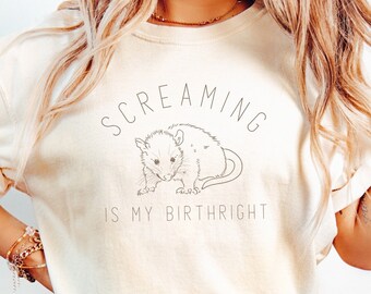 May include: Ivory-colored t-shirt with the text "SCREAMING IS MY BIRTHRIGHT" above a line drawing of an opossum. The shirt is paired with blue jeans.