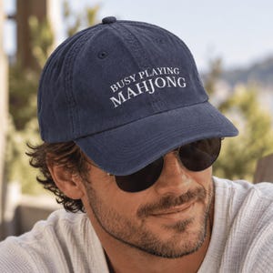 Puede incluir: Una gorra de béisbol azul marino con el texto bordado en blanco BUSY PLAYING MAHJONG en el panel frontal. La gorra tiene una visera curva, un botón en la parte superior y ojales cosidos para la ventilación, hecha de tela suave y texturizada.