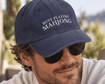 May include: A navy blue baseball cap featuring the white embroidered text BUSY PLAYING MAHJONG on the front panel. The cap has a curved brim, a button on the top, and stitched eyelets for ventilation, constructed from a soft, textured fabric.