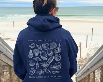 May include: A navy blue hoodie with a white graphic of seashells and the text "where your treasure is there will be your heart also". The hoodie is being worn by a person standing on a wooden boardwalk overlooking a beach.