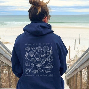 Christian girl contemplating faith staring at the ocean while wearing her navy blue gildan beachy hoodie with white seashells and bible verse that says, "where your treasure is there will be your heart also."