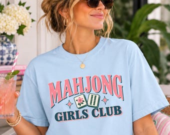 May include: A light blue short-sleeved t-shirt featuring the text "MAHJONG GIRLS CLUB" in pink and dark green lettering. The design includes a graphic of two mahjong tiles with floral and bamboo patterns, accented by small four-pointed stars.
