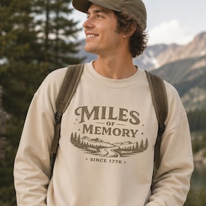 Puede incluir: Una sudadera de cuello redondo color crema con un estampado gráfico marrón oscuro en el pecho. El diseño incluye el texto "MILES OF MEMORY" sobre una ilustración de una carretera sinuosa, pinos y montañas, con el texto "SINCE 1776" debajo.