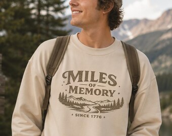 May include: A cream-colored crewneck sweatshirt featuring a dark brown graphic print on the chest. The design includes the text "MILES OF MEMORY" above an illustration of a winding road, pine trees, and mountains, with the text "SINCE 1776" below.