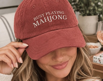 May include: A rust-colored corduroy baseball cap featuring the embroidered white text BUSY PLAYING MAHJONG across the front panel. The hat has a structured crown, a curved brim, and stitched eyelets for ventilation on the top panels.