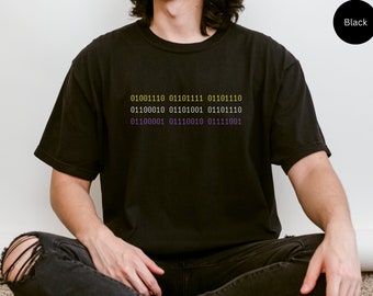 Puede incluir: Una camiseta negra con un estampado gráfico de código binario en amarillo y morado. El código dice: 01001110 01101111 01101110 01100010 01101001 01101110 01100001 01110010 01111001