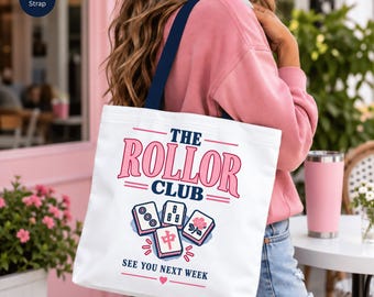 May include: A white canvas tote bag with a navy blue strap featuring the text THE ROLLOR CLUB and SEE YOU NEXT WEEK in pink and navy blue. The design includes four mahjong tiles with pink and navy blue floral and character illustrations.