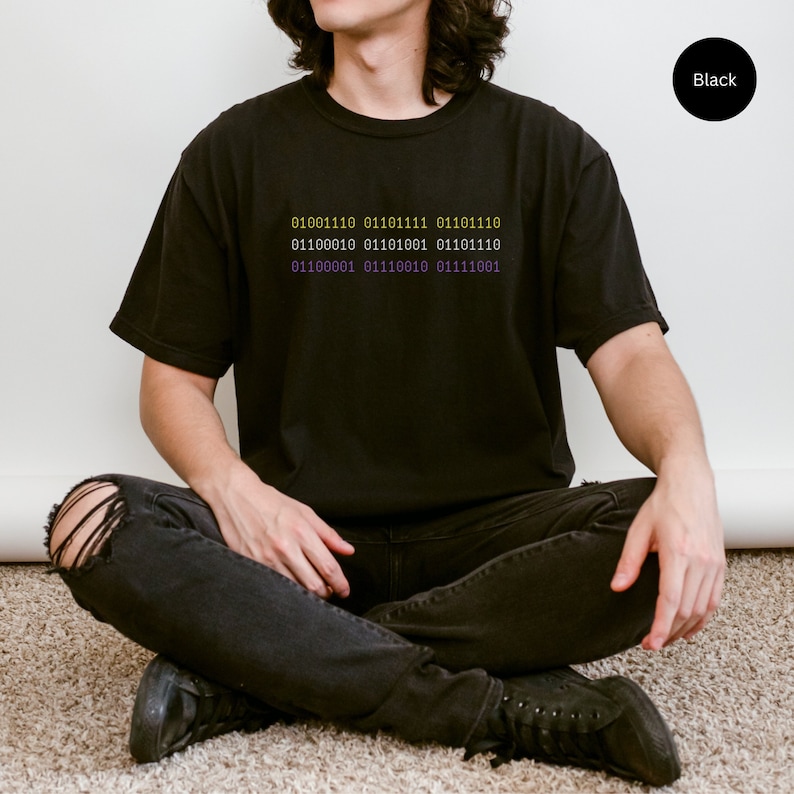 Agender nonbinary person wearing futuristic techwear black Comfort Colors t-shirt that says "nonbinary" in nonbinary flag colors using binary code for an ironic shirt twist.