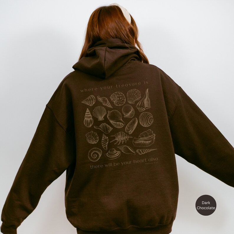 Puede incluir: Una sudadera con capucha de color marr&oacute;n chocolate oscuro con un estampado gr&aacute;fico de conchas marinas y el texto "where your treasure is there will be your heart also".