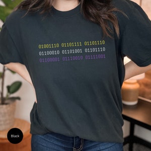Puede incluir: Una camiseta negra con un dise&ntilde;o de c&oacute;digo binario morado y amarillo. El c&oacute;digo dice: 01001110 01110111 01101110 01100010 01101001 01101110 01100001 01110010 01111001