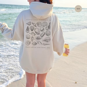 Puede incluir: Una sudadera con capucha de color crema con un gr&aacute;fico de conchas marinas y el texto "where your treasure is there will be your heart also Matthew 6:21" en la parte posterior.