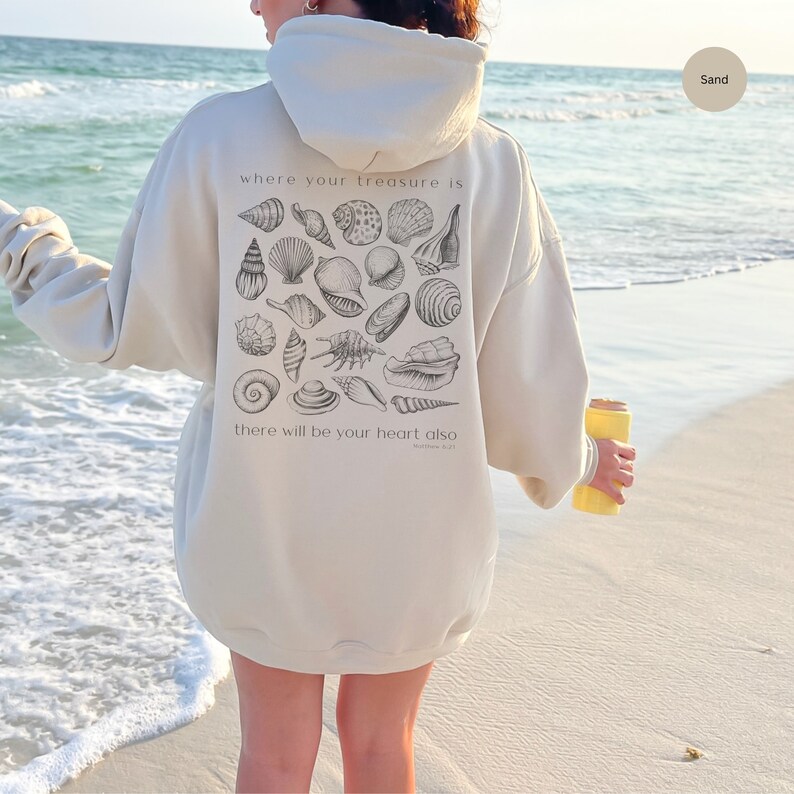 Puede incluir: Una sudadera con capucha de color crema con un gr&aacute;fico de conchas marinas y el texto "where your treasure is there will be your heart also Matthew 6:21" en la parte posterior.