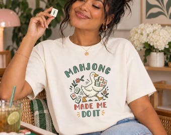 May include: An off-white short-sleeve t-shirt featuring a graphic of a white goose carrying mahjong tiles. The text above the goose reads "MAHJONG" in green, and the text below reads "MADE ME DO IT" in red, surrounded by small floral accents.