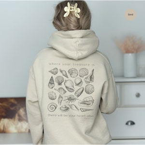 Puede incluir: Una sudadera con capucha beige con un gr&aacute;fico de conchas marinas y el texto "where your treasure is there will be your heart also Matthew 6:21" impreso en la espalda.