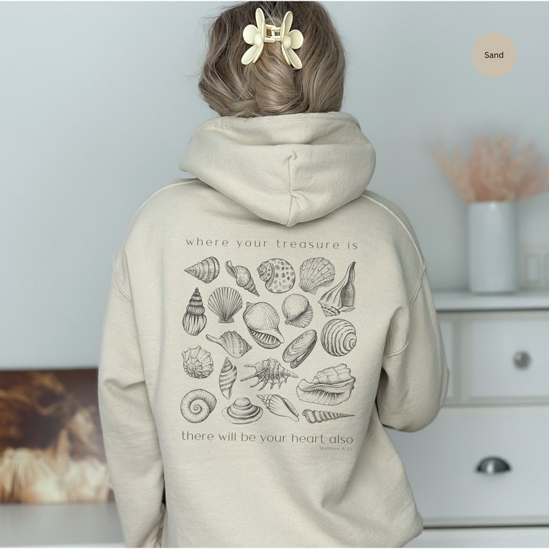 Puede incluir: Una sudadera con capucha beige con un gr&aacute;fico de conchas marinas y el texto "where your treasure is there will be your heart also Matthew 6:21" impreso en la espalda.