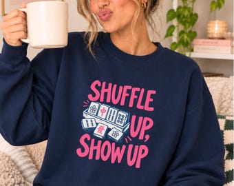 May include: A navy blue crewneck sweatshirt featuring a graphic of mahjong tiles and the pink text "SHUFFLE UP, SHOW UP". The design includes a set of tiles arranged in a tray, with one tile displaying a red Chinese character and others showing floral patterns.
