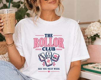 May include: A white short-sleeve t-shirt featuring a graphic with the text THE ROLLOR CLUB in pink and navy blue. Below the text are four mahjong tiles in pink, white, and navy blue, followed by the text SEE YOU NEXT WEEK and a small pink heart.