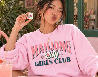 Puede incluir: Una sudadera de cuello redondo rosa claro con el texto MAHJONG GIRLS CLUB en letras rosas y verde oscuro, acompañada de ilustraciones de dos fichas de mahjong, una con un diseño de flor rosa y la otra con marcas de bambú verdes.