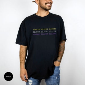 Puede incluir: Una camiseta negra con un gr&aacute;fico de c&oacute;digo binario en amarillo y morado. El c&oacute;digo dice: "01001110 01101111 01101110 01100010 01101001 01101110 01100001 01110010 01111001".