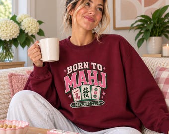 May include: A maroon crewneck sweatshirt featuring the text BORN TO MAHJ in pink and white letters, with three mahjong tiles and the text MAHJONG CLUB in green and white. The design includes small pink star accents on a dark red fabric background.