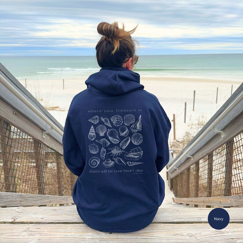 Christian girl contemplating faith staring at the ocean while wearing her navy blue gildan beachy hoodie with white seashells and bible verse that says, "where your treasure is there will be your heart also."