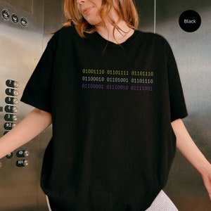 Puede incluir: Camiseta negra con un gr&aacute;fico de c&oacute;digo binario en amarillo y morado. El c&oacute;digo dice: "01001110 01101111 01101110 01100010 01101001 01101110 01100001 01110010 01111001".