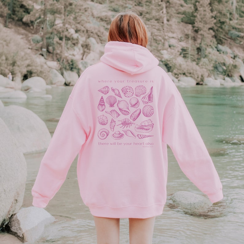 Puede incluir: Sudadera rosa con un gr&aacute;fico de conchas marinas y la cita "Donde est&eacute; tu tesoro, all&iacute; estar&aacute; tambi&eacute;n tu coraz&oacute;n." La cita es de Mateo 6:21.