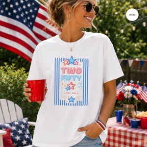 Puede incluir: Camiseta blanca con la inscripción "TWO FIFTY" en azul y celeste, con estrellas y los años 1776-2026. La camiseta se lleva al aire libre, con una bandera estadounidense al fondo.