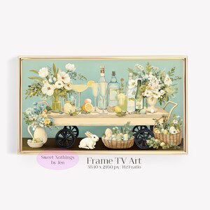 May include: A framed digital art print featuring a decorative cart with floral arrangements, bottles, glasses, lemons, and Easter eggs. A white bunny sits near a basket of eggs. The art has a light blue background and gold frame. Text reads "Sweet Nothings by Jen" and "Frame TV Art".
